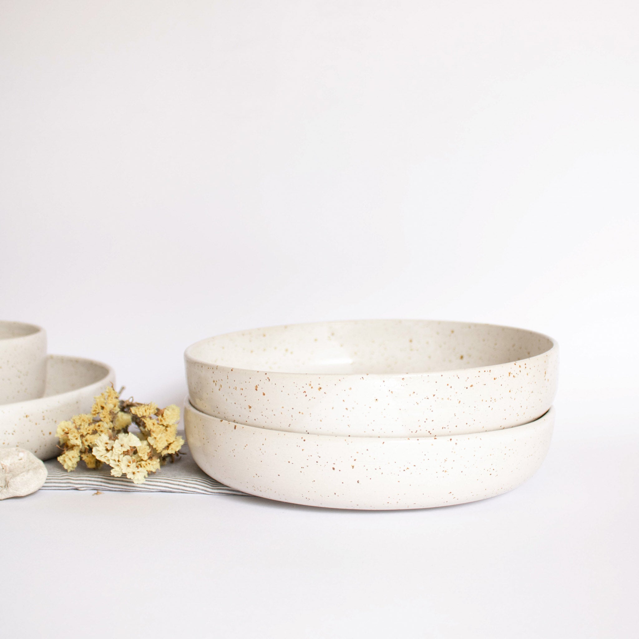 Handcrafted ceramic pasta plates in speckled white finish, displayed alongside dry flowers on a white background.