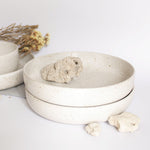 Handcrafted ceramic pasta plates in speckled white finish, displayed alongside rocks and dry flowers on a white background.