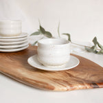 Handcrafted ceramic Ripple cup and saucer plate in speckled white finish, displayed on a wooden board.