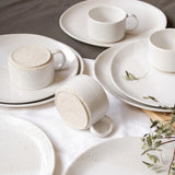 Artfully scattered on a table, speckled white handmade ceramic cups with handles rest atop matching speckled white plates.