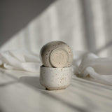 A stack of two handcrafted espresso cups in speckled white finish, with the top one featuring a maker's mark detail at the bottom of the cup.