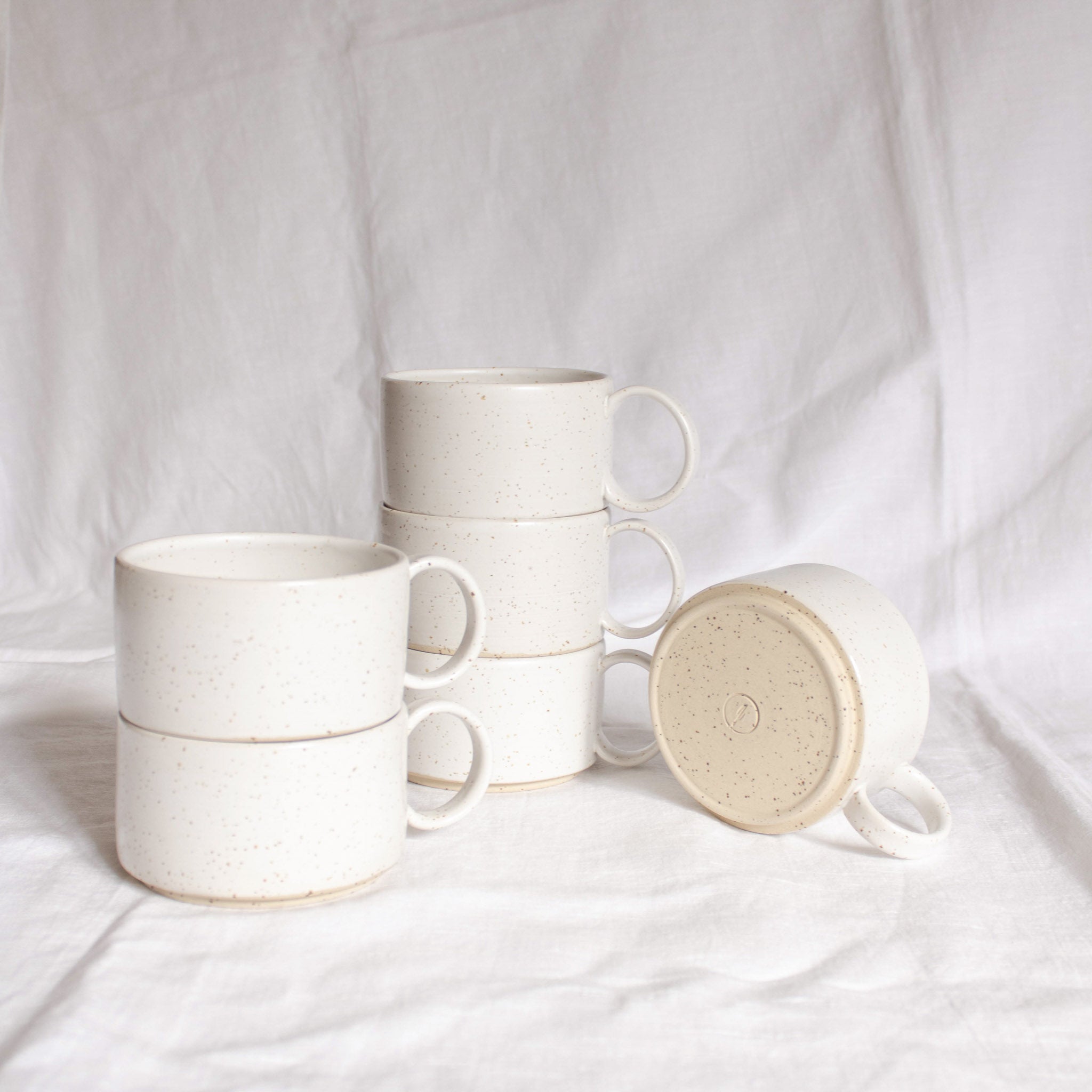 A group of handmade ceramic cups with handles, made from speckled cream clay, glazed in satin white glaze, presented against a white backdrop.