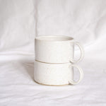 A stack of two ceramic cups with handles, made from speckled cream clay, glazed in satin white glaze, presented against a white backdrop.