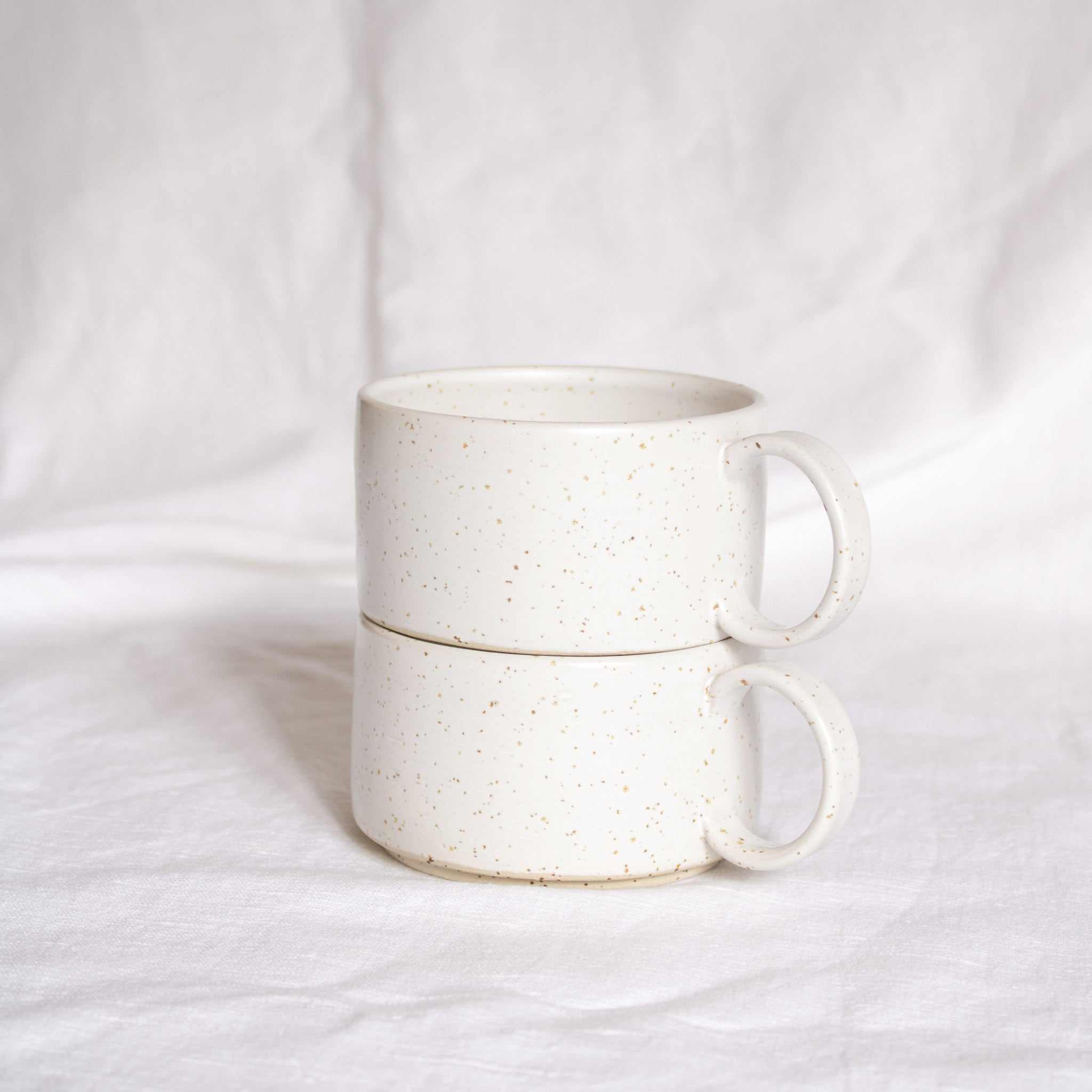 A stack of two ceramic cups with handles, made from speckled cream clay, glazed in satin white glaze, presented against a white backdrop.