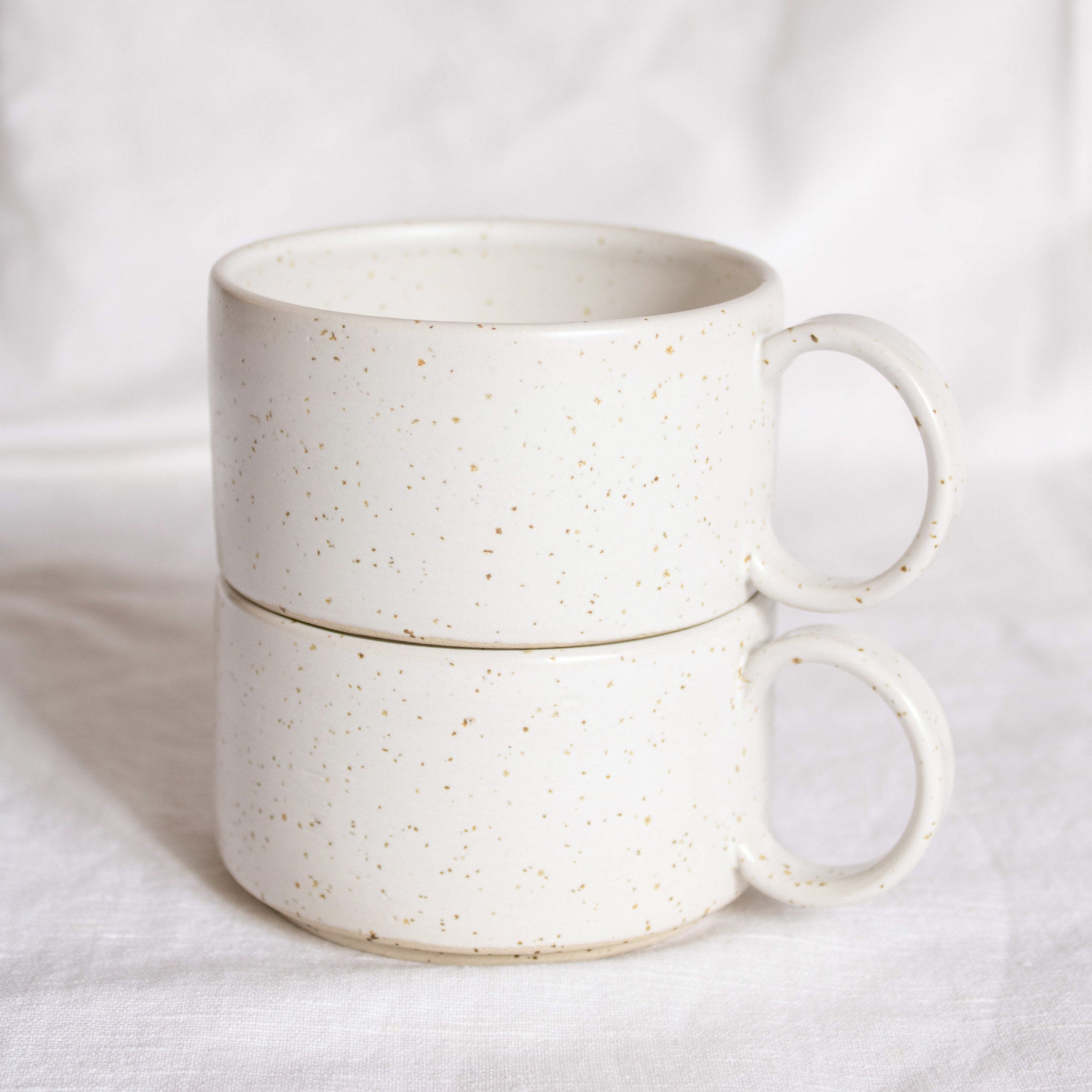 Close-up of two ceramic cups with handles, made from speckled cream clay, glazed in satin white glaze, presented against a white backdrop.