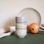 A stack of cappuccino cups with a matte marble finish in natural earthy tones, displayed alongside draped fabric and fruits.