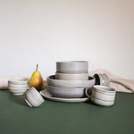 Handmade ceramic pasta plates and cups in a matte earthy finish, arranged on a table, accompanied by fruits and drapery.