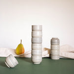 Stacks of espresso cups with a matte marble finish in natural earthy tones, displayed alongside draped fabric and fruits.