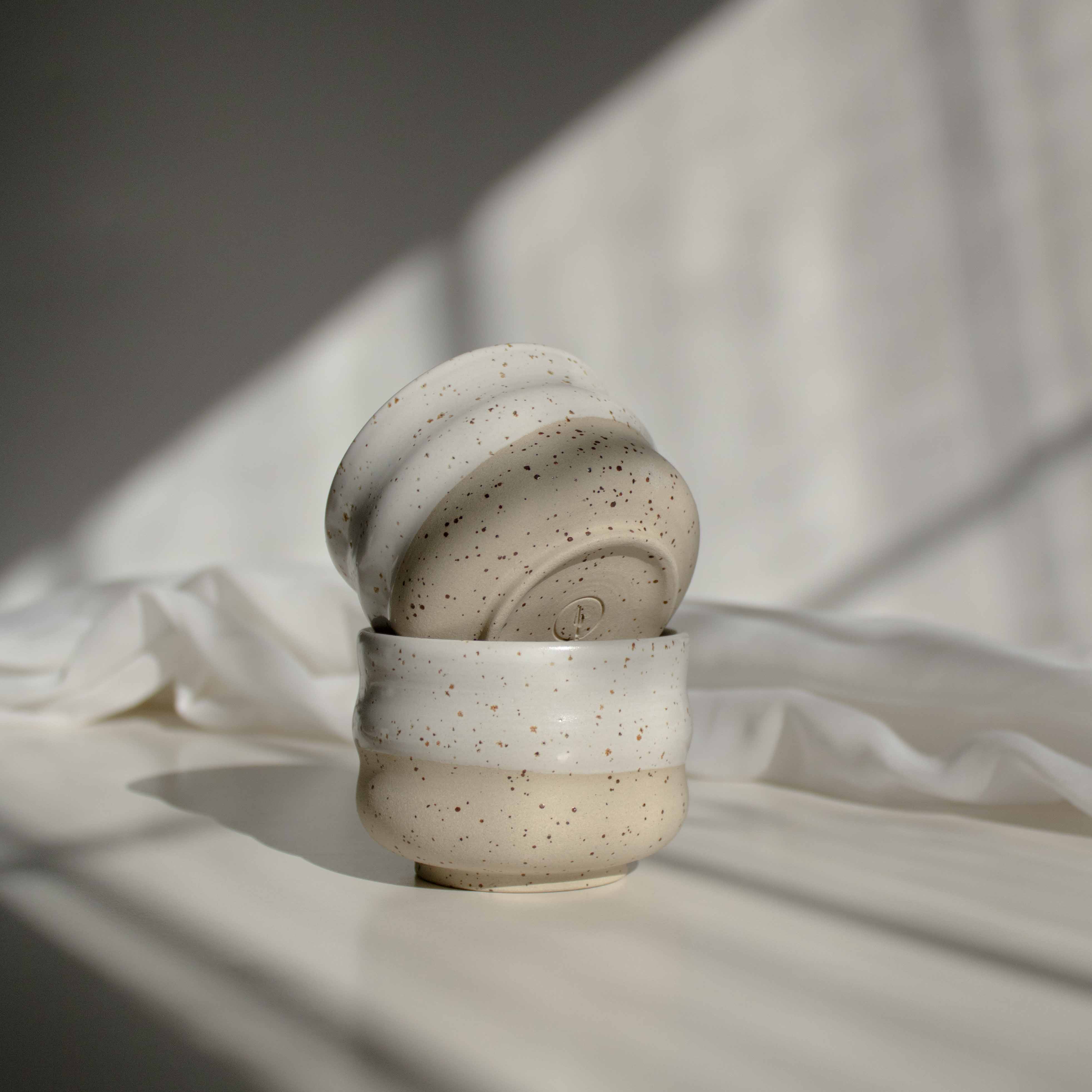 A stack of two handcrafted cappuccino cups in speckled white finish, with the top one featuring a maker's mark detail at the bottom of the cup.