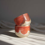 Two handcrafted cappuccino cups in coral red finish, stacked and illuminated by contrasting light and shadows.