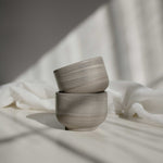 Two handcrafted cappuccino cups in earthy tones, stacked and illuminated by contrasting light and shadows.