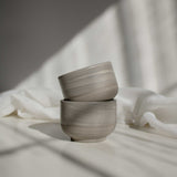 Two handcrafted cappuccino cups in earthy tones, stacked and illuminated by contrasting light and shadows.