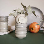 A stack of cappuccino cups with a matte marble finish in natural earthy tones, displayed alongside draped fabric and fruits.