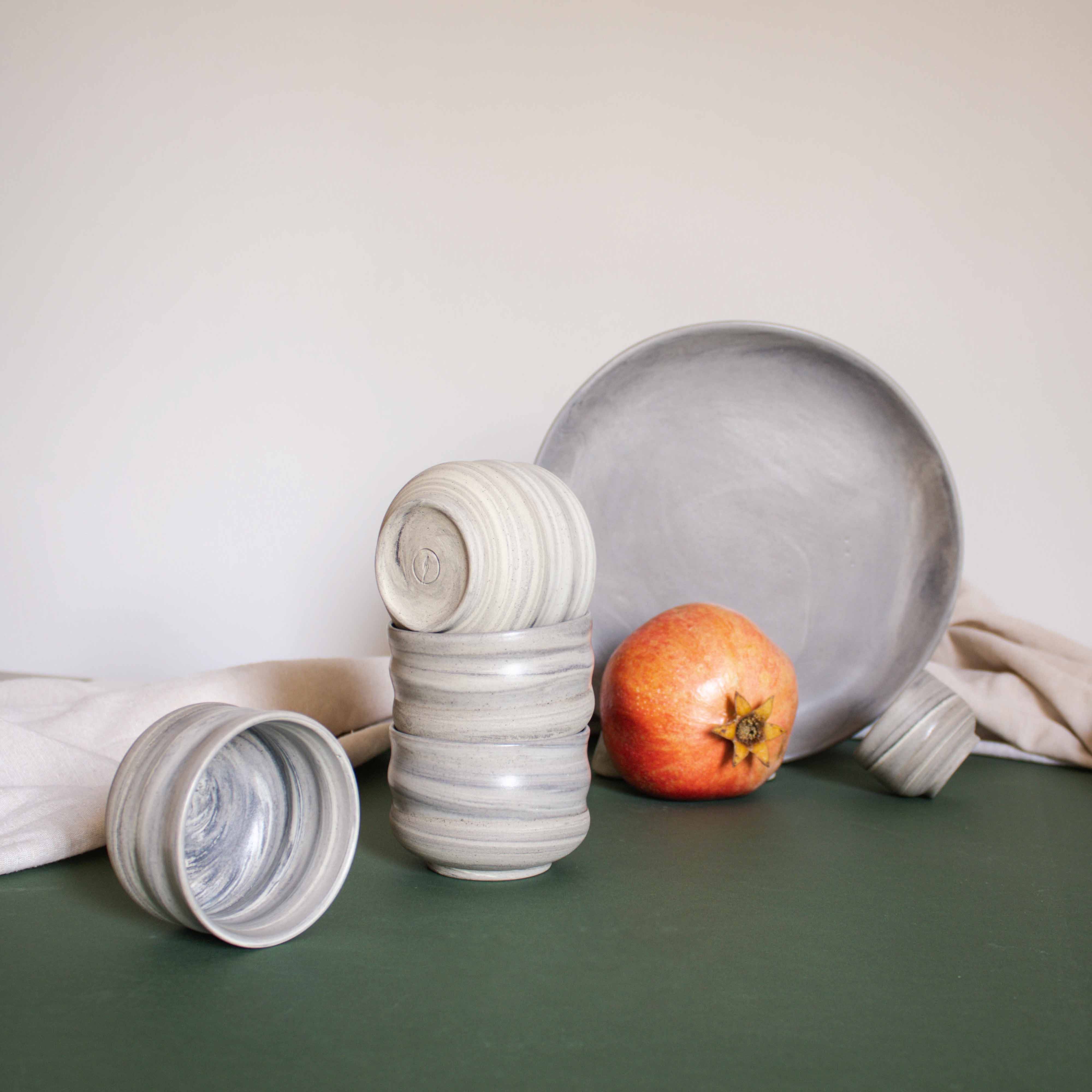 A stack of cappuccino cups with a matte marble finish in natural earthy tones, displayed alongside draped fabric and fruits.