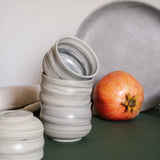 A stack of cappuccino cups with a matte marble finish in natural earthy tones, displayed alongside draped fabric and fruits.