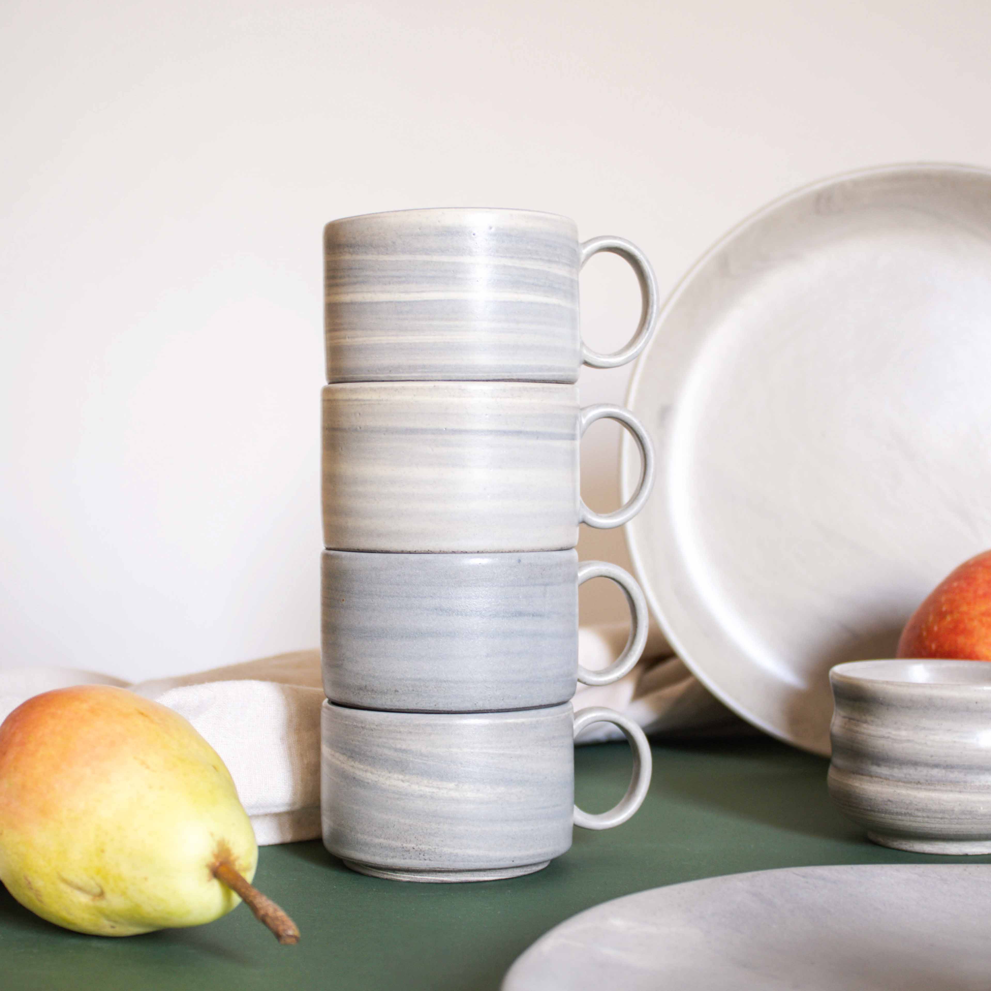 
An arrangement of artisanal ceramic cups with handles, neatly stacked on a tabletop alongside other tableware and fruits.